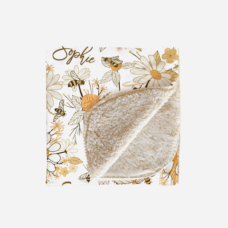 May include: A white throw blanket featuring a repeating pattern of yellow and brown bees, daisies, and wildflowers. The name Sophie is printed in brown script in the upper left corner. The blanket has a folded corner revealing a tan sherpa lining.