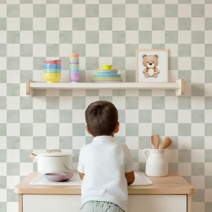 Peut inclure: Un enfant se tient devant un comptoir en bois avec un pot en céramique blanche, une cuillère en bois et un petit bol violet. Le comptoir est devant un mur avec un motif à carreaux vert et blanc. Une étagère au-dessus du comptoir contient une pile de bols colorés, une pile d'assiettes colorées et un cadre avec une image d'un ours en peluche brun.
