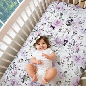 May include: A crib sheet featuring a white background with a floral pattern of light purple and dark violet roses, small buds, and green leaves. The sheet is fitted over a mattress inside a light-colored wooden crib with vertical slats.