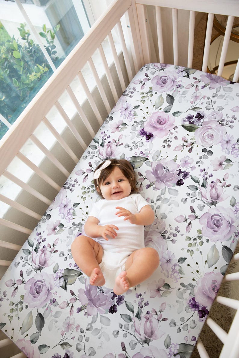 May include: A crib sheet featuring a white background with a floral pattern of light purple and dark violet roses, small buds, and green leaves. The sheet is fitted over a mattress inside a light-colored wooden crib with vertical slats.