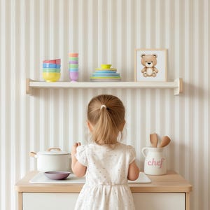 May include: A young child stands in front of a white kitchen counter with a white pot, a purple bowl, and a white ceramic jar with the word "chef" written in pink. The child is wearing a white dress with a floral pattern. The counter is against a striped wallpaper background.