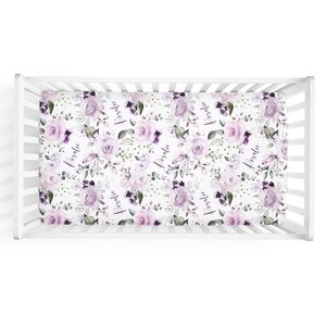 May include: A white crib sheet featuring a repeating pattern of light purple and dark violet roses, green leaves, and the name Rosalie printed in a black cursive script, fitted onto a white wooden crib with vertical slats.