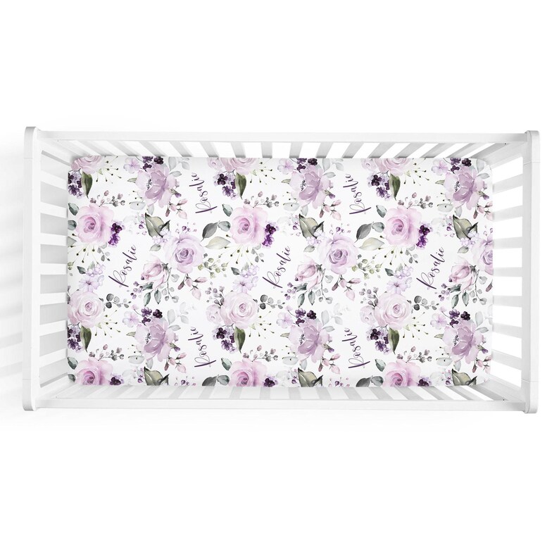 May include: A white crib sheet featuring a repeating pattern of light purple and dark violet roses, green leaves, and the name Rosalie printed in a black cursive script, fitted onto a white wooden crib with vertical slats.
