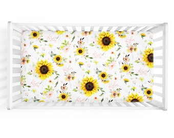 May include: White crib sheet with a sunflower and floral pattern. The sheet has the name "Kyla" repeated throughout the design.