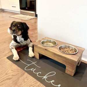 May include: A black and white dog sits on a gray mat with the name "Tucker" written in white. The dog is looking at a wooden raised dog bowl stand with two stainless steel bowls. One bowl is filled with water and the other with dog food.