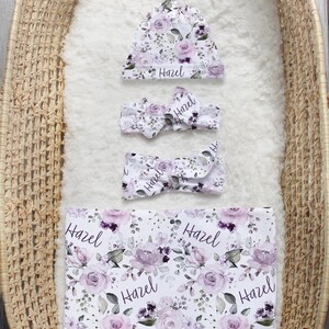 May include: A white baby set featuring a floral pattern of purple roses and green leaves with the name Hazel printed in black script. The set includes a knotted hat, two headbands with bows, and a rectangular swaddle blanket.