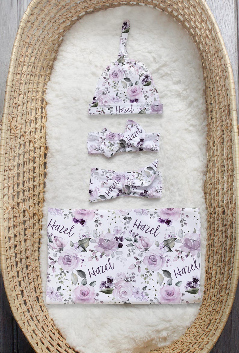 May include: A white baby set featuring a floral pattern of purple roses and green leaves with the name Hazel printed in black script. The set includes a knotted hat, two headbands with bows, and a rectangular swaddle blanket.
