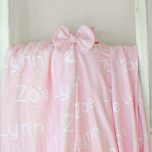 May include: A pink baby blanket with the names "Zoe" and "Lynn" printed in white on it. A white bow is on top of the blanket.