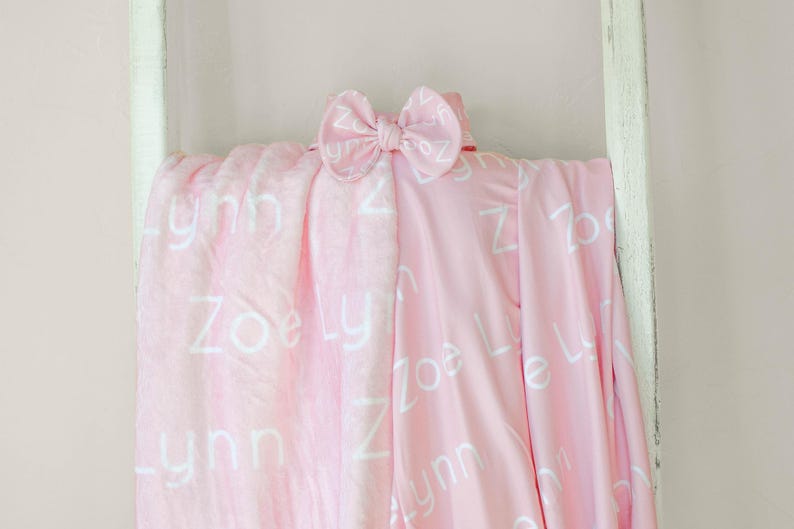 May include: A pink baby blanket with the names "Zoe" and "Lynn" printed in white on it. A white bow is on top of the blanket.