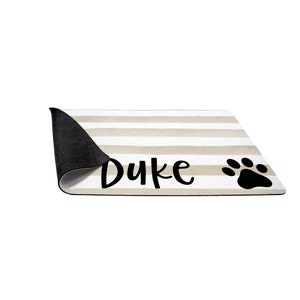 May include: A beige and white striped dog doormat with black text that says "Duke" and a black paw print.