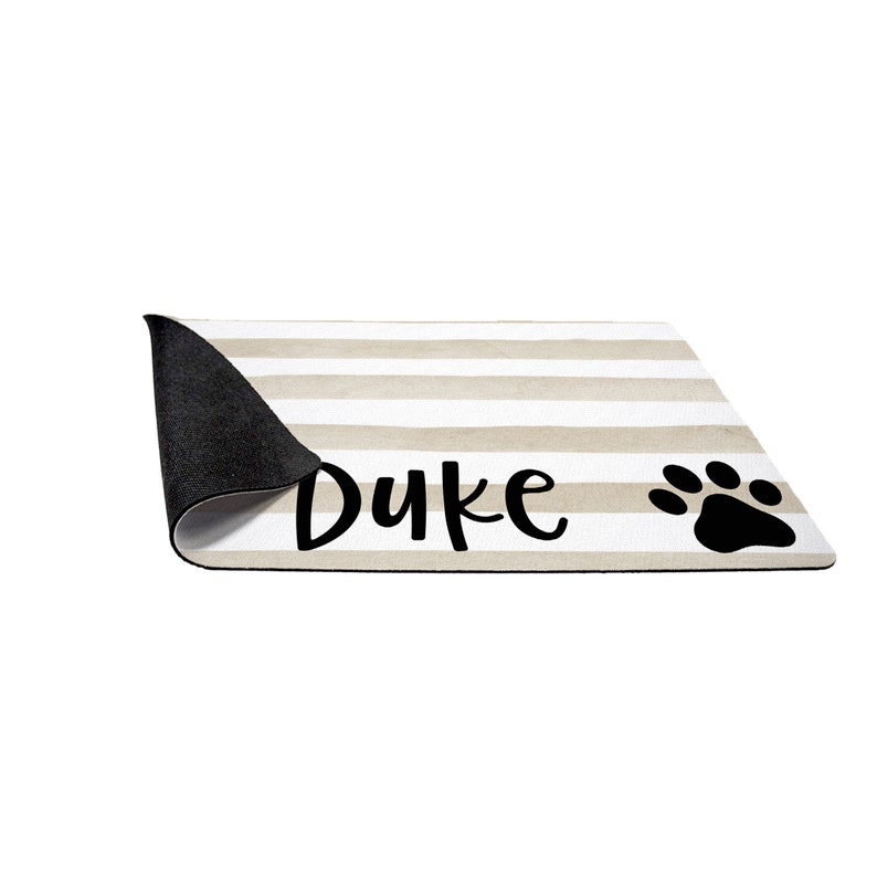 May include: A beige and white striped dog doormat with black text that says "Duke" and a black paw print.