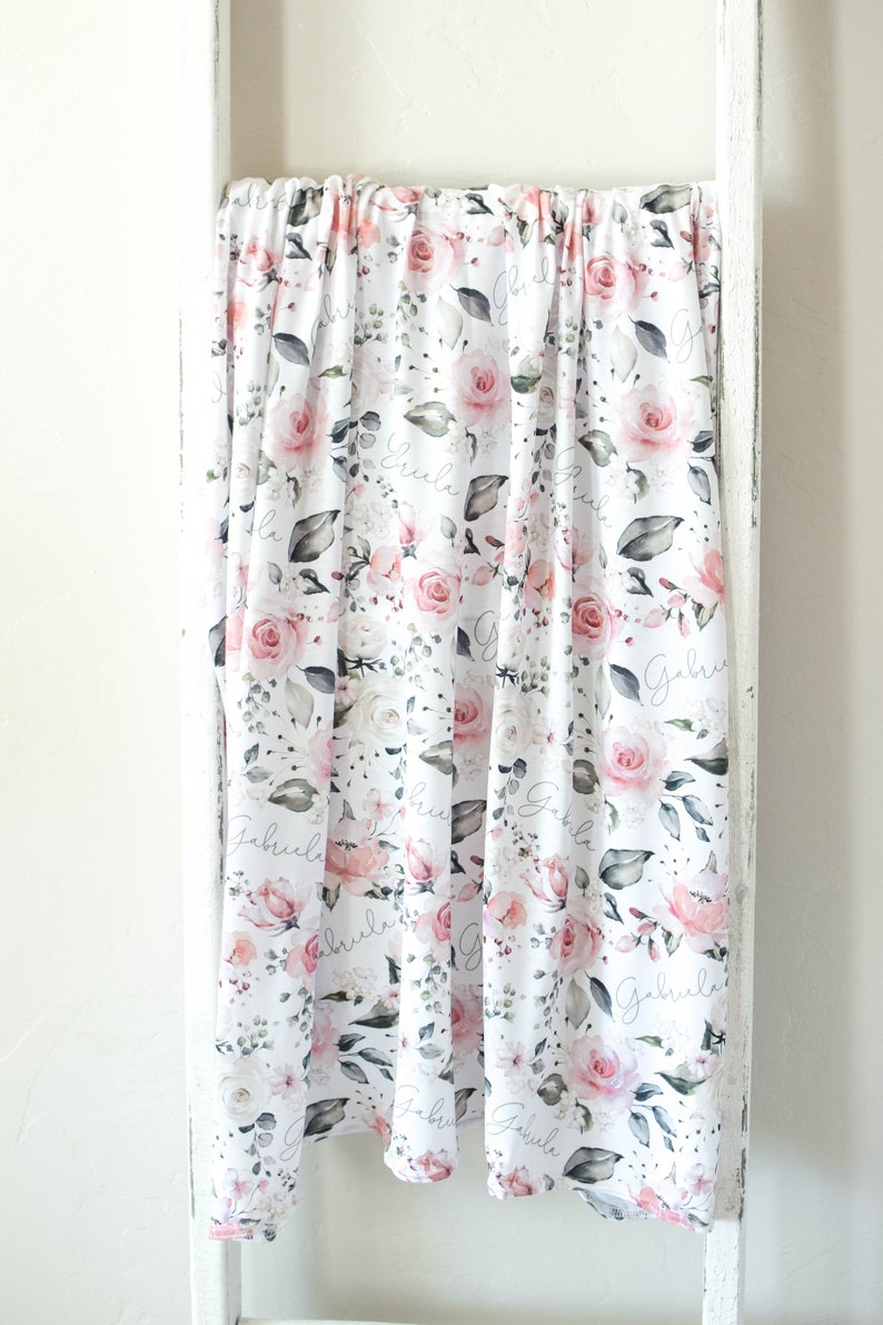 K&ouml;nnte beinhalten: Eine wei&szlig;e Musselin-Wickeldecke mit einem rosafarbenen Blumenmuster und dem Namen "Gabriella", der sich &uuml;ber das gesamte Design wiederholt.