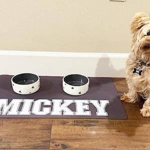 May include: A brown dog food mat with the name "MICKEY" printed in white. Two white ceramic dog bowls with black spots are on the mat. A small, light brown dog is sitting on the floor next to the mat.