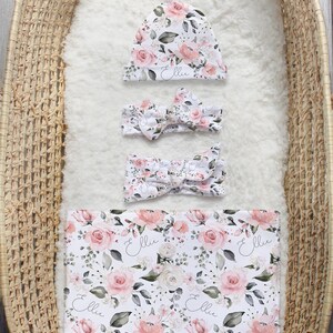 K&ouml;nnte beinhalten: Ein florales Set aus M&uuml;tze, Stirnband und Wickeltuch f&uuml;r ein Baby M&auml;dchen. Das Set hat ein rosa-wei&szlig;es Blumenmuster mit dem Namen "Ellie" in Kursivschrift auf jedem Teil.