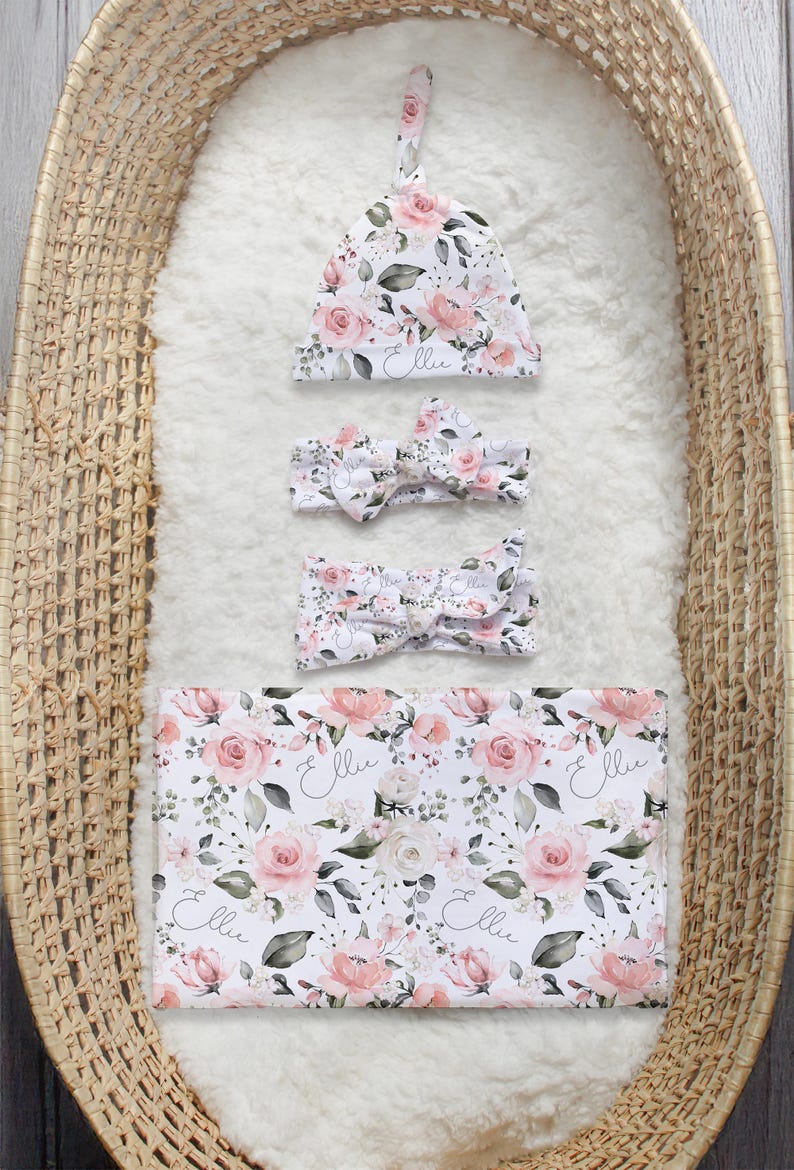 K&ouml;nnte beinhalten: Ein florales Set aus M&uuml;tze, Stirnband und Wickeltuch f&uuml;r ein Baby M&auml;dchen. Das Set hat ein rosa-wei&szlig;es Blumenmuster mit dem Namen "Ellie" in Kursivschrift auf jedem Teil.
