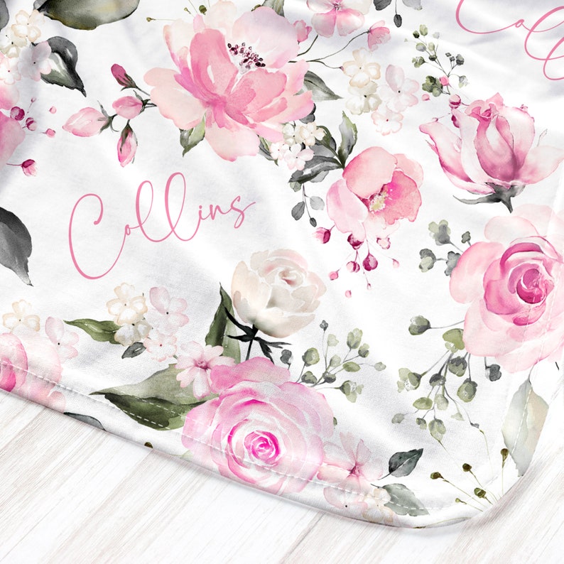 May include: White blanket with a watercolor floral design featuring pink roses and other flowers. The word "Collins" is written in pink script across the blanket. Green leaves and accents complete the design.