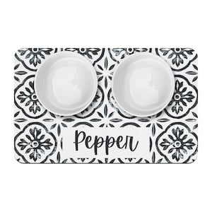 May include: White ceramic pet food bowls with a black and white patterned mat. The mat has the name "Pepper" printed on it.