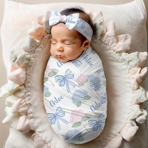 May include: A newborn baby wrapped in a white swaddle blanket and wearing a matching headband, both adorned with blue bows, pink and blue hydrangea flowers, and the name "Chloe". The baby is resting on a ruffled pillow.