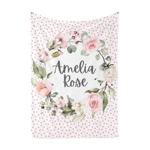Puede incluir: Una manta de beb&eacute; blanca con un estampado de lunares rosas. El centro presenta una corona floral con el nombre "Amelia Rose" en escritura cursiva. La corona incluye rosas rosas, vegetaci&oacute;n y otras flores.