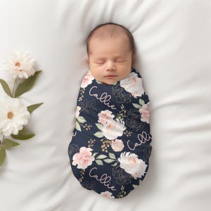 May include: A newborn baby swaddled in a navy blue blanket with a floral design. The blanket features pink and white roses, gold accents, and the name "Callie" in a delicate script. The baby is lying on a white surface.