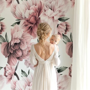 May include: A woman in a white dress holds a baby in front of a wall mural. The mural features large, watercolour-style pink and mauve peony flowers with green leaves against a white background. A white curtain is visible on the right.