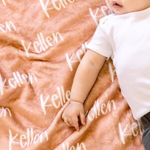 May include: A soft, peach-colored blanket with the name "Kellen" written in white script across the fabric. A baby is lying on the blanket.