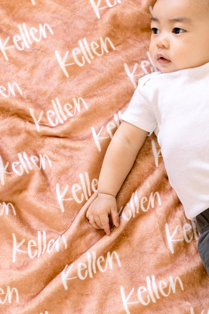 May include: A soft, peach-colored blanket with the name "Kellen" written in white script across the fabric. A baby is lying on the blanket.