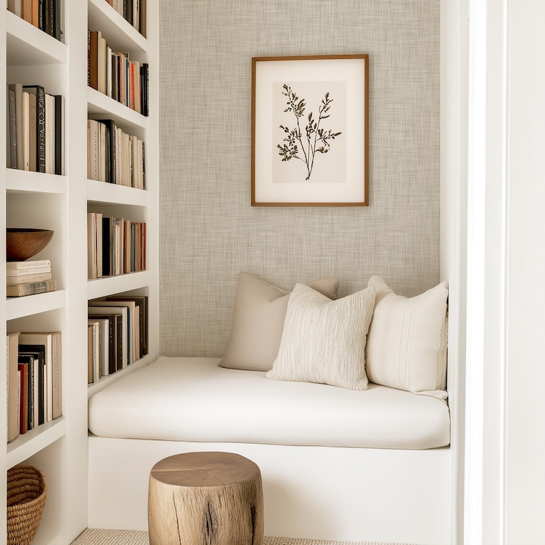 May include: A white built-in bench with a white cushion and three throw pillows. The bench is in a nook with white built-in shelves filled with books. A wooden stool sits in front of the bench. A framed print of a botanical illustration hangs on the wall.