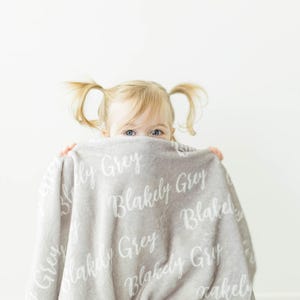 May include: A young child hides behind a soft, gray blanket with the name "Blakely Grey" printed on it in white.