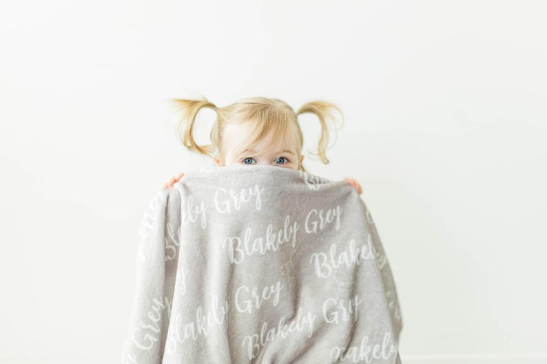 May include: A young child hides behind a soft, gray blanket with the name "Blakely Grey" printed on it in white.