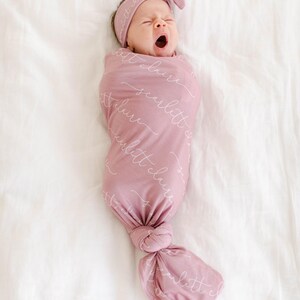 May include: A pink swaddle blanket with the text "little cloud" repeated in white script. A baby is swaddled in the blanket and is wearing a pink headband.
