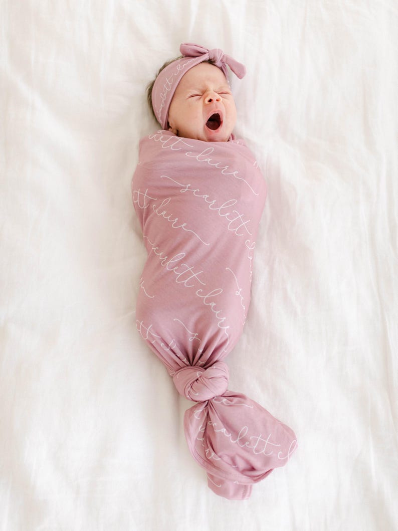 May include: A pink swaddle blanket with the text "little cloud" repeated in white script. A baby is swaddled in the blanket and is wearing a pink headband.