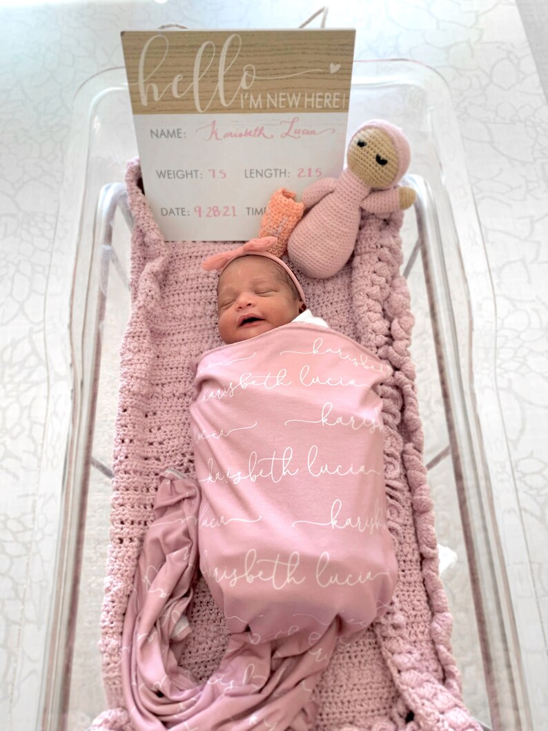 May include: A newborn baby girl swaddled in a pink blanket with the name "Karysbeth Lucia" written in white script. The baby is wearing a pink headband and is lying in a clear plastic bassinet. A pink crocheted doll is lying next to the baby. A wooden sign with the text "hello I'M NEW HERE" is above the bassinet. The sign also includes the baby's name, weight (7.5 lbs), length (21.5 inches), and date of birth (9-28-21).