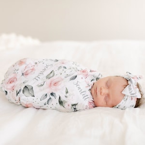 May include: A sleeping baby wrapped in a white swaddle with a pink floral pattern and the name "Scarlett" written on it. The baby is wearing a matching pink floral headband with the word "Sweet" written on it.
