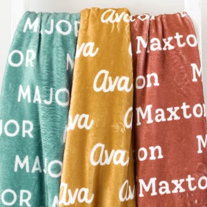 May include: Three personalized fleece blankets with the names "Major", "Avalon", and "Maxto" printed in white on a teal, mustard yellow, and rust colored background.