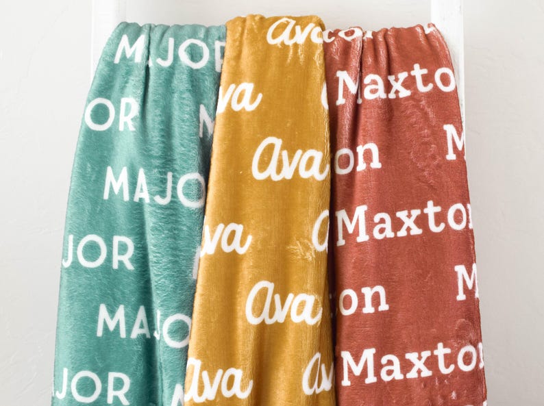 May include: Three personalized fleece blankets with the names "Major", "Avalon", and "Maxto" printed in white on a teal, mustard yellow, and rust colored background.