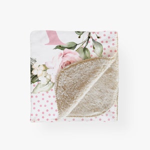 Puede incluir: Una manta blanca con un estampado de lunares rosas y un dise&ntilde;o floral. El dise&ntilde;o floral incluye rosas rosas, flores blancas y hojas verdes. La manta tiene un suave forro de sherpa beige.