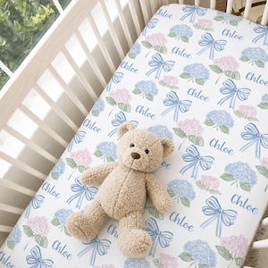 May include: A white crib sheet featuring a repeating pattern of blue and pink hydrangea flowers, light blue bows, and the name Chloe in a cursive script, with a tan plush teddy bear resting on top of the fabric inside a white wooden crib.