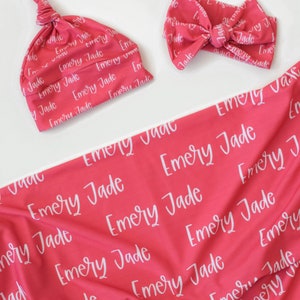 May include: A pink baby blanket and matching hat and headband with the name "Emery Jade" printed in white on the fabric.