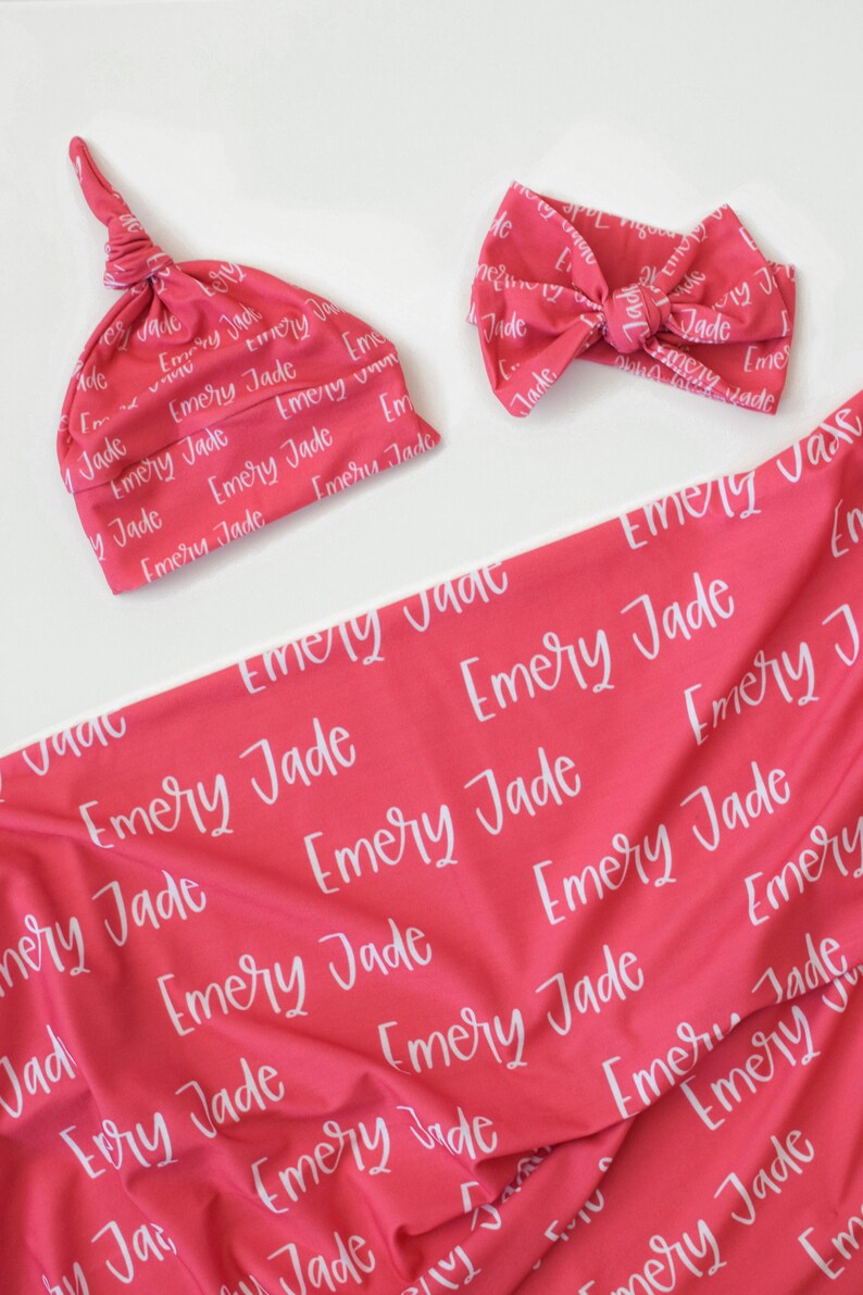 May include: A pink baby blanket and matching hat and headband with the name "Emery Jade" printed in white on the fabric.