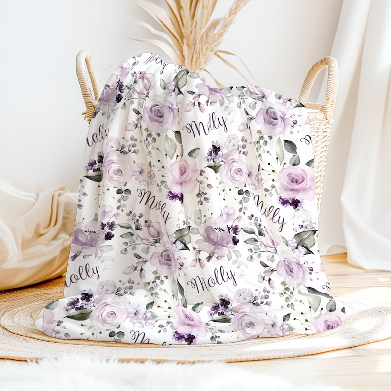 May include: A white throw blanket featuring a repeating pattern of light purple roses, dark purple floral accents, and green leaves. The name Molly is printed in a black cursive script throughout the fabric, which is draped over a woven basket.