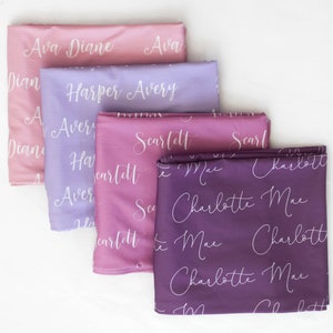 May include: Four fabric squares with different names written in white script on each. The squares are stacked on top of each other, with the top square being a light pink colour, the second square being a light purple colour, the third square being a darker pink colour, and the bottom square being a dark purple colour. The names on the squares are Ava Diane, Harper Avery, Scarlett, and Charlotte Mae.