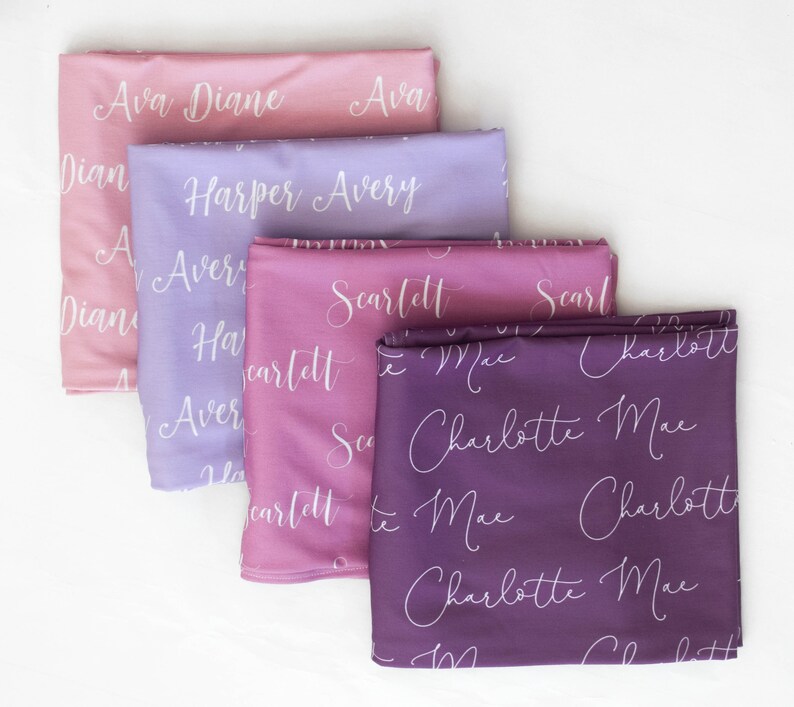 May include: Four fabric squares with different names written in white script on each. The squares are stacked on top of each other, with the top square being a light pink colour, the second square being a light purple colour, the third square being a darker pink colour, and the bottom square being a dark purple colour. The names on the squares are Ava Diane, Harper Avery, Scarlett, and Charlotte Mae.