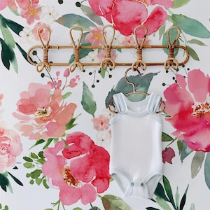 May include: A white baby onesie with thin straps hangs on a wooden rack against a backdrop of a watercolor floral wallpaper featuring pink and green flowers.