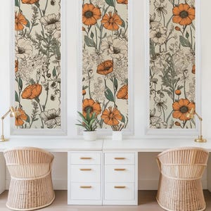 May include: Three framed prints with a floral pattern featuring orange flowers and green leaves on a cream background. The prints are hanging on a white wall above a white desk with two wicker chairs and drawers.