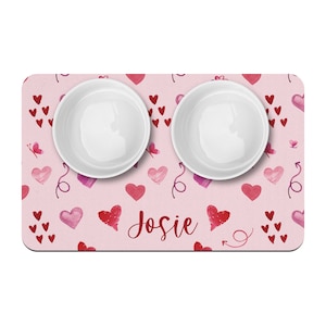 May include: Pink pet food mat with a heart pattern and the name "Josie" printed on it. Two white ceramic bowls are placed on the mat.