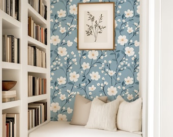May include: A blue wallpaper featuring a repeating pattern of white flowers with dark green stems and leaves. The wallpaper is applied to a wall behind a white built-in bench with three beige throw pillows and a framed botanical print.