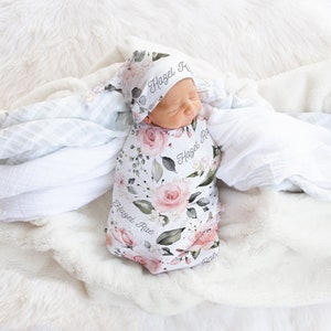 K&ouml;nnte beinhalten: Ein wei&szlig;er Swaddle mit floralem Muster und dem Namen "Hazel Rae" darauf gedruckt. Ein Baby tr&auml;gt den Swaddle und eine passende florale M&uuml;tze mit dem Namen "Hazel Rae" darauf gedruckt.