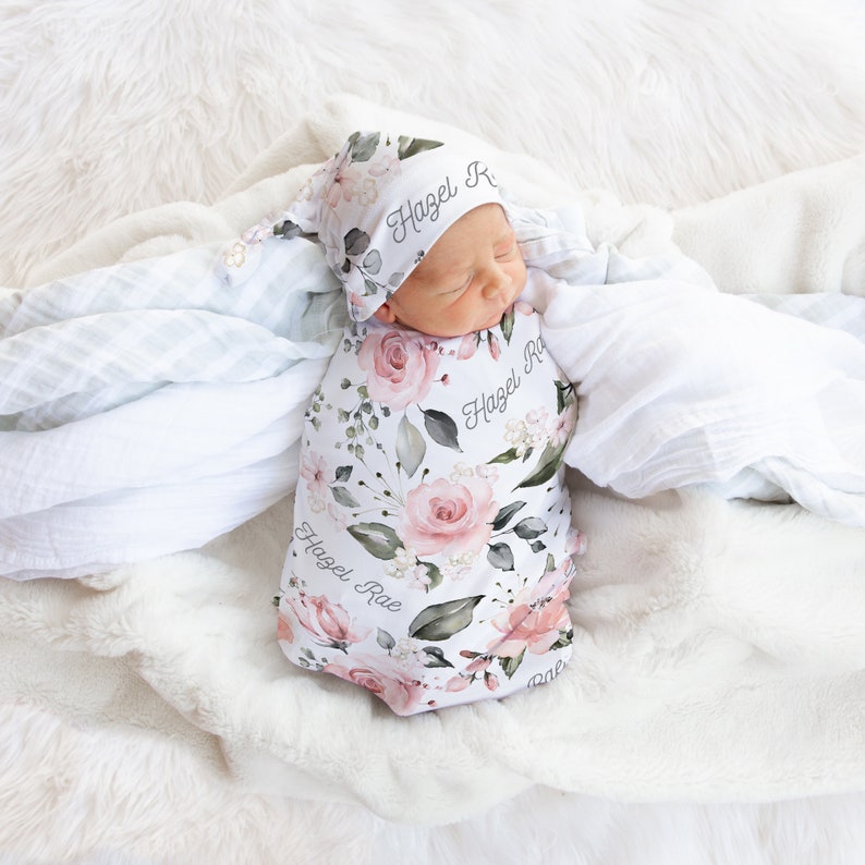 K&ouml;nnte beinhalten: Ein wei&szlig;er Swaddle mit floralem Muster und dem Namen "Hazel Rae" darauf gedruckt. Ein Baby tr&auml;gt den Swaddle und eine passende florale M&uuml;tze mit dem Namen "Hazel Rae" darauf gedruckt.