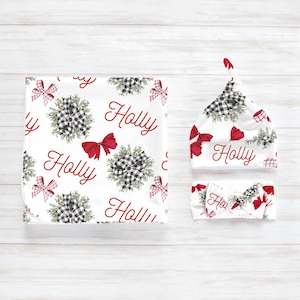 May include: A white baby blanket, hat, and headband set. The set features a repeating pattern of black and white buffalo check bows, red bows, and greenery. The word "Holly" is written in red script across the items.